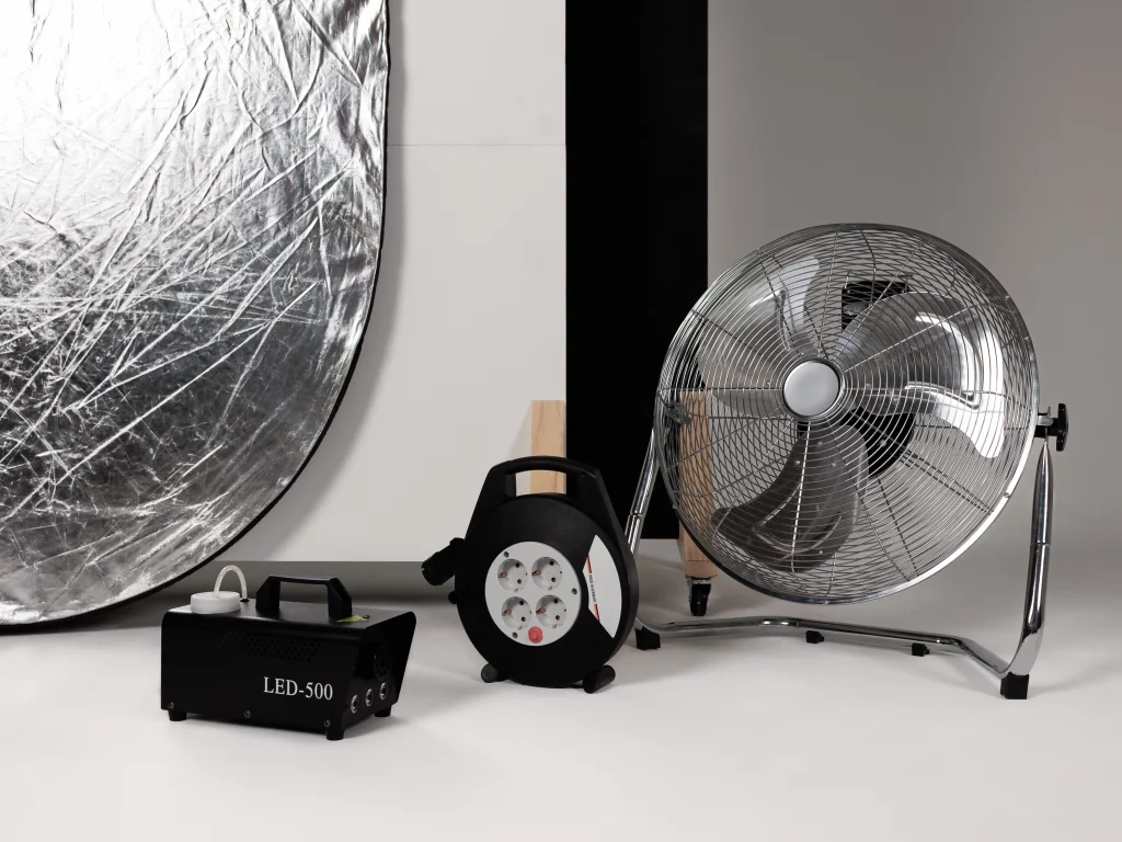Fotostudio-Ausrüstung mit silbernem Reflektor, einer LED‑500 Effektmaschine, Kabeltrommel und verchromtem Bodenventilator auf weißem Hintergrund.